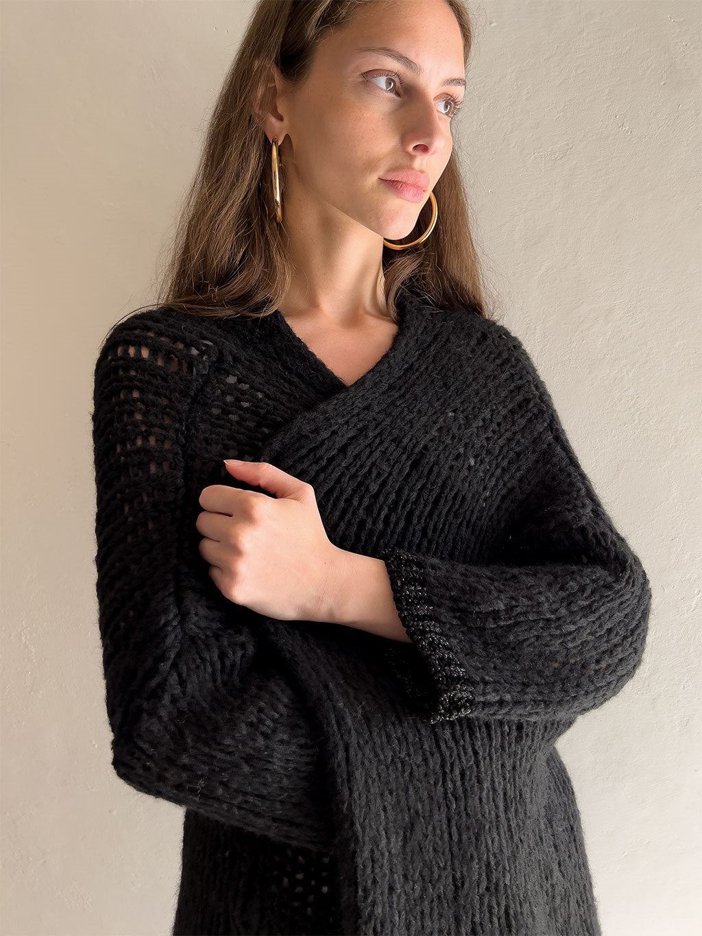 A person with long, brown hair is facing the camera, wearing a black KYLA CARDIGAN by DEBBIE KATZ over a white top and white pants. The background is plain and light-colored. They have a neutral expression and natural makeup.