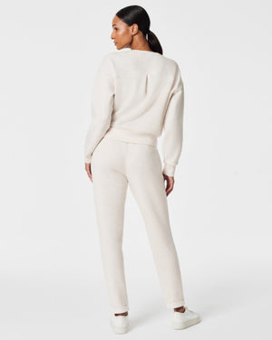 A person stands against a white background, smiling and looking down with one hand touching their hair. They are wearing a matching light beige SPANX AIRESSENTIALS TAPERED PANT and sweatshirt set made from ultra drapey, lightweight spacer fabric, paired with white sneakers.