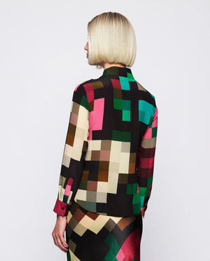 A woman with short blonde hair is wearing the MIRTO 1956 - MULTICOLOR SILK PLAID BLOUSE paired with a matching skirt. The chic fashion outfit from MIRTO 1956 features a collar and buttons down the front, showcasing green, pink, black, and brown squares on a white background.