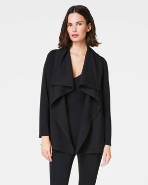 A person wearing the SPANX - AIRESSENTIALS DRAPE FRONT CARDIGAN in black, designed with long sleeves and crafted from lightweight fabric, elegantly poses with their hand in one of its pockets. The cardigan features an asymmetrical hem and is paired beautifully with black pants, highlighting the side and pocket details against a plain background.