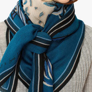 Close-up of an INOUI EDITIONS - WESTERN SQUARE SCARF featuring a blue and beige abstract pattern. The 100% wool fabric appears soft and slightly wavy, showcasing intricate weaving and a smooth texture. The unique design includes shades of blue, black, and beige with curved and dotted shapes. Perfect for adding a stylish touch to any outfit.