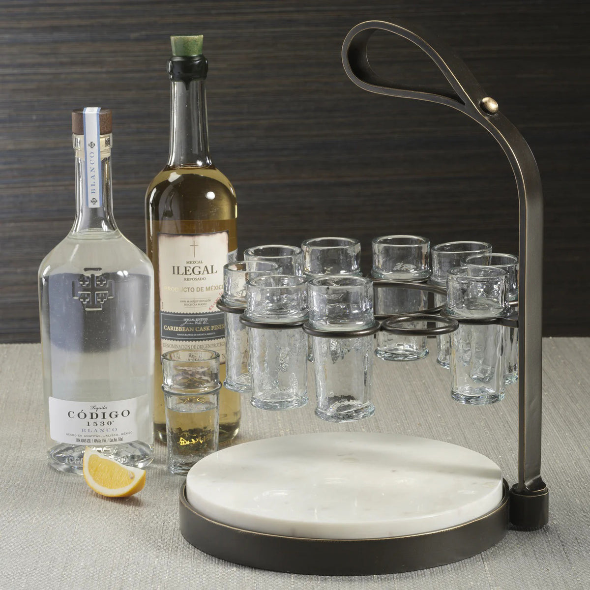 The ZODAX TEQUILERO 10 Shot Tequila Set features a circular white marble cutting board with an iron handle and metal frame, holding ten empty shot glasses arranged in a ring.