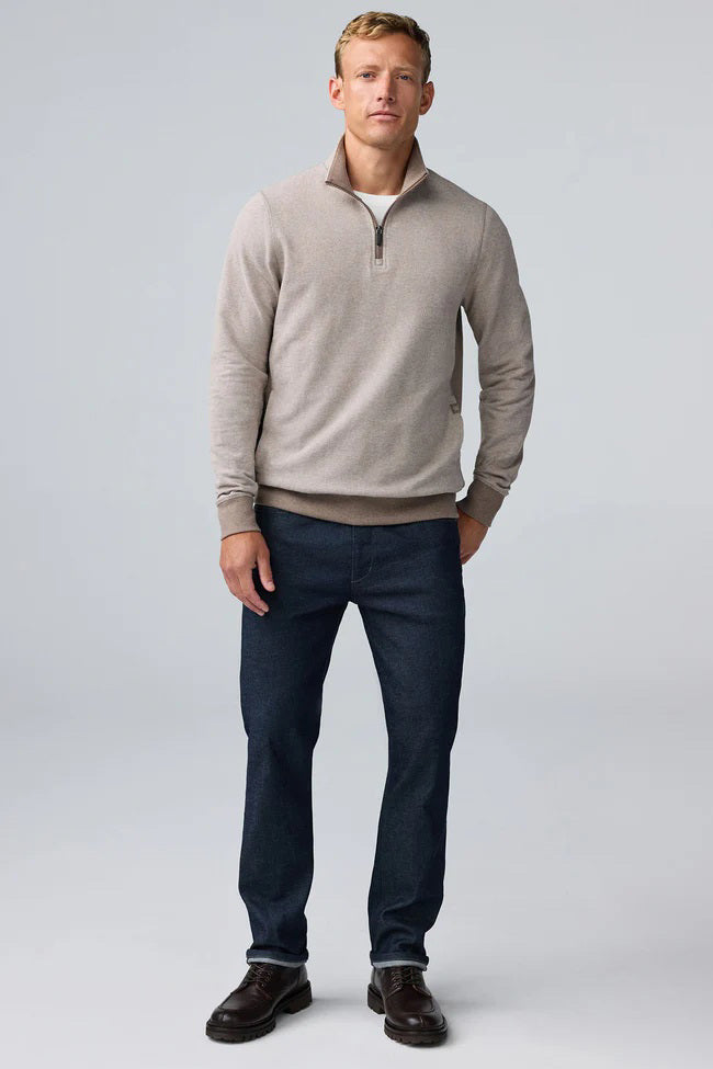 A man with short light brown hair stands in profile, wearing the MACK WELDON Ace 1/2 Zip Sweater in Herringbone and dark pants, one hand in his pocket, against a plain light gray background.