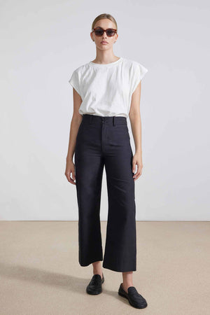 In a minimalist setting, an individual confidently poses against a neutral background, wearing sleek sunglasses, a white T-shirt, the APIECE APART - CLASSIC MERIDA PANT in black high-waisted wide-legged style, and stylish black loafers.