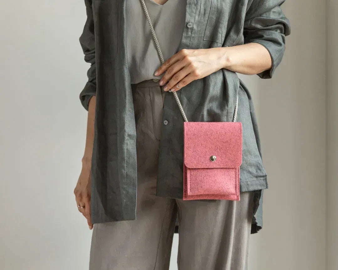 A person in a beige sweater and tan pants holds the WOOLBERRY WOOL FELT PHONE BAG, a small gray handcrafted bag with a flap and button closure. The image is cropped at the shoulders and knees.