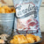 A light blue bag with a rustic wooden plank design labeled "CAROLINA KETTLE - CRAB BOIL CHIPS 5OZ" from the brand "1 IN 6 SNACKS". Featuring a red crab holding a map, the text "Certified Awesome" and "Guaranteed Fressher," 5 oz (141.8g), GF, and the "1 in 6 Snacks" logos. Enjoy this savory treat inspired by Southern tradition.