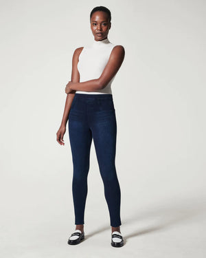 A person stands confidently in front of a plain background. They are wearing a white sleeveless turtleneck top, SPANX - JEANNISH ANKLE LEGGINGS in dark blue, and black and white loafers. Their arms are relaxed, and they gaze directly into the camera.