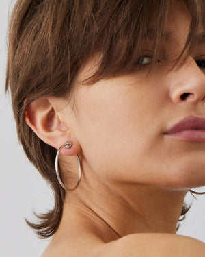 Two JENNY BIRD SMALL ICON HOOP EARRINGS crafted from stainless steel lie on a light gray surface. Featuring a minimalist design with a spherical accent, the clean backdrop highlights their sleek elegance and timeless appeal.