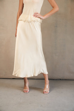 A person wearing a navy blue SATIN MIDI SKIRT from ROSE & CROWN paired with a matching navy blue top. The individual is also wearing beige pointed-toe heels. The background features a plain, light-colored wall with a concrete floor.