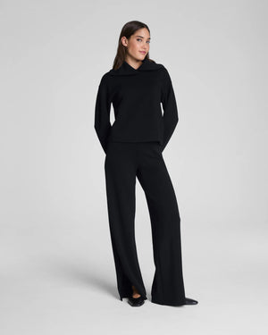 A person stands with one hand in their pocket, wearing SPANX AirEssentials Zero Waistband Wide Leg Pant made from light-as-air fabric and pointed black shoes against a plain light background.