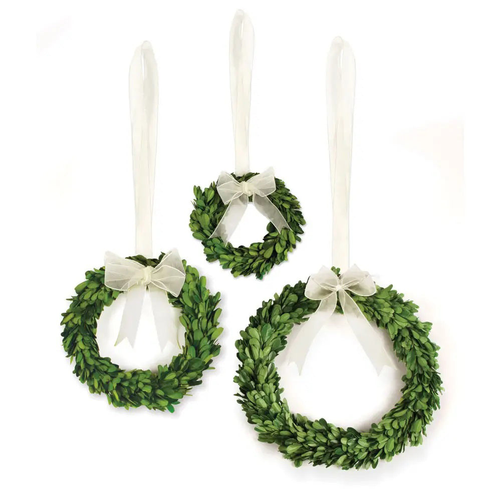 A large NAPA HOME AND GARDEN preserved boxwood wreath, featuring a sheer white ribbon artfully tied into a bow at the top, is displayed against a plain background.