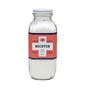 A clear glass bottle labeled "BAUDELAIRE - RECOVER BATH SELTZER 18OZ" with a red and white label. The label highlights that it is handmade, therapeutic, features an effervescent formula, contains all-natural essential oils, and weighs 18 ounces.