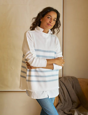 A woman with long dark hair wears a Monterey Rolled Funnel Neck Stripe Sweater from FRANK & EILEEN. The sweater, made of machine-washable Italian cotton, features light gray stripes on the back. She layers it over a white shirt and gazes to the side in a softly lit neutrally toned room.