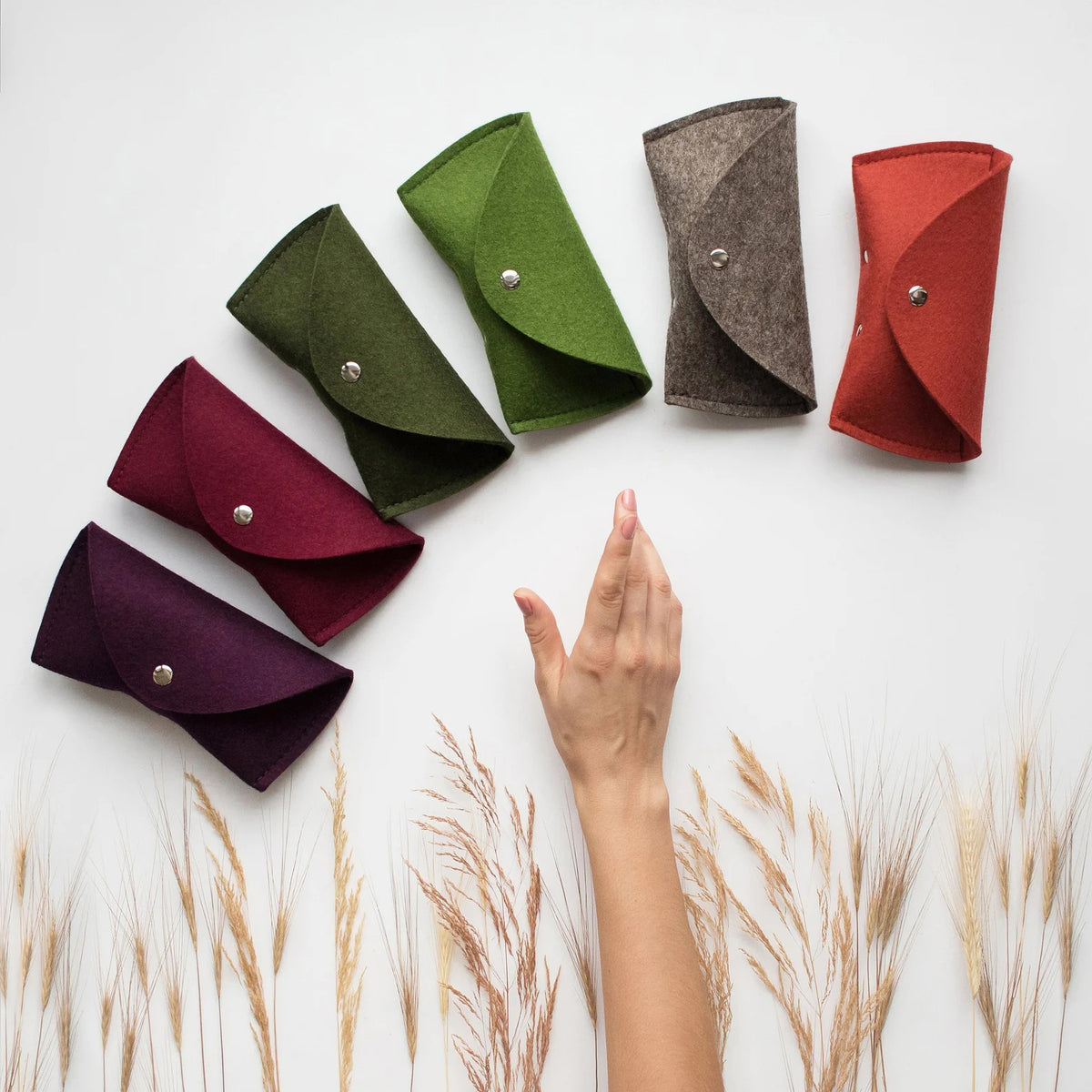 Three WOOLBERRY WOOL FELT GLASSES CASES in navy, bright, and light blue with magnetic closures are arranged on a white surface near clear sunglasses, framed by white-blue hydrangeas.