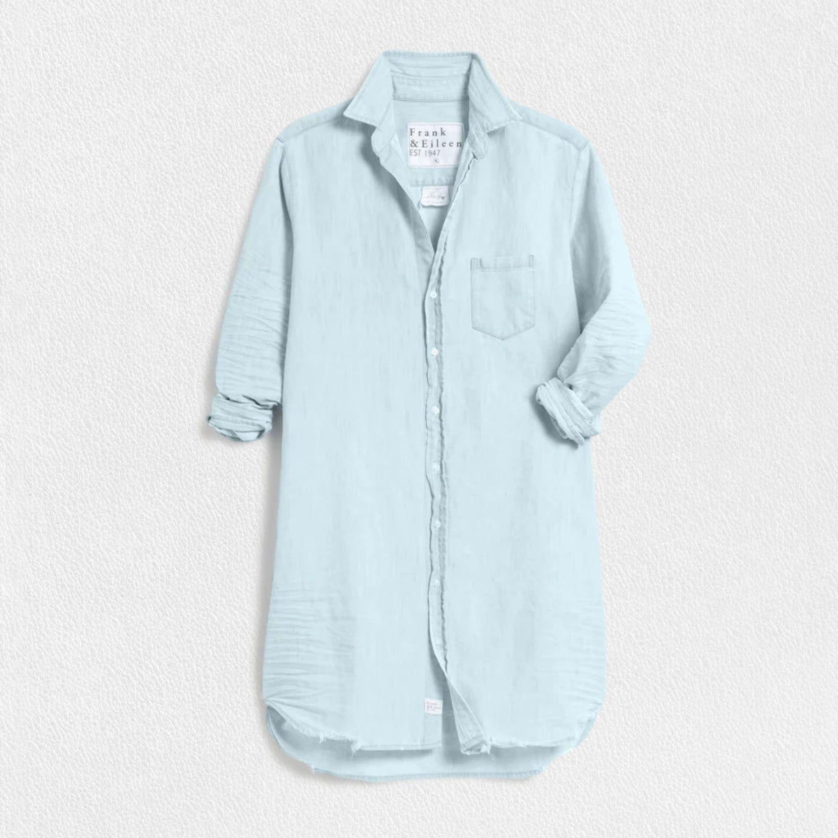 The FRANK AND EILEEN - MARY CLASSIC SHIRTDRESS in Classic Blue Tattered Wash by FRANK &amp; EILEEN is a light blue, long linen shirtdress featuring a button-down front and long sleeves rolled up to three-quarter length. It has a single chest pocket and resembles the casual elegance of Famous Denim with its textured fabric and slightly frayed hem, displayed against a textured white background.