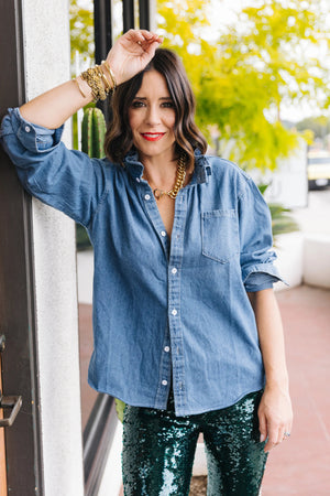 A woman is pictured from the shoulders down, wearing the versatile KMJ - QUARANTINA SHIRT, a white button-up with rolled-up sleeves, paired with blue jeans. She has her hands in her pockets and accessorizes with layered gold necklaces and bracelets. Her nails are manicured and painted white.