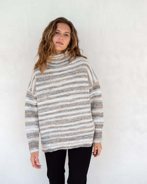 A person wearing the MERSEA - GOOD VIBRATIONS STRIPE TURTLENECK from MER SEA, a cozy sweater made of nylon, polyester, and acrylic in shades of white, gray, and beige, leans against a countertop. They have curly hair and a relaxed posture. The background includes a white vase and several stacked items.