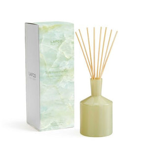 The LAFCO - Wild Honeysuckle Diffuser set features a green, angular glass bottle with wooden reeds inserted. The accompanying packaging, placed next to the bottle, showcases a light green and white marbled design with the LAFCO logo and scent description, accentuating the floral fragrance notes of this essential oil-based creation.