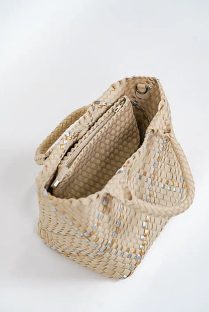 A person in a beige sleeveless top and ripped blue jeans holds a Parker & Hyde - Beige Metallic Woven Tote Bag, featuring a checkered design with subtle white accents and two handles. The background is plain light gray.