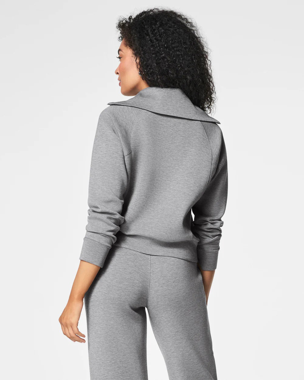 A woman with curly hair stands with her back to the camera, enveloped in the SPANZ - AIRESSENTIALS HALF ZIP tracksuit by SPANX. The top features a wide collar and long sleeves, while the pants are fitted. Crafted from an ultra-light fabric, this ensemble effortlessly combines comfort and style against a plain white backdrop.