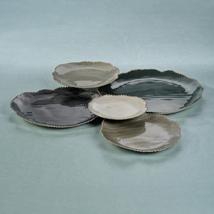 The ZODAX ENAMELED 5 CLUSTER SERVEWARE SET features five decorative plates arranged on a white surface. Each plate varies in size and displays shades of black and grey. The irregular edges are adorned with gold trim, giving the set an organic, leaf-like appearance.