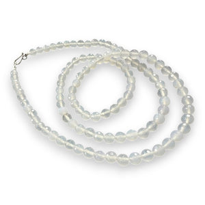 A coiled ANN LIGHTFOOT - LONG STRAND FACETED MILKY AGATE NECKLACE of translucent, round beads is displayed on a dark wooden surface. The faceted 8mm beads have a smooth and glossy finish, appearing luminous against the dark background. The necklace is attached with a sterling silver clasp.