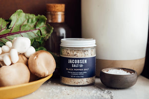 The JACOBSEN SALT - INFUSED BLACK PEPPER SALT is housed in a glass jar with a white lid. The label lists sea salt and black pepper as the ingredients and notes the absence of anti-caking agents, so natural clumping may occur. It asserts that the product is not bioengineered and touts its suitability for enhancing recipes. This product is manufactured by JACOBSEN SALT CO. in Portland, OR.