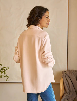 A woman with dark, wavy hair faces left wearing the FRANK & EILEEN Eileen Sweatshirt Button Up in Petal and blue jeans. She stands in a softly lit room with a beige backdrop, a draped chair behind her, and some greenery to her left.