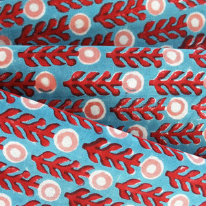 Close-up of the CHRISTINA DICKSON HOME - POPPY COTTON NAPKIN, handmade in India with 100% cotton. Features red leaf-like shapes and white circular motifs with pink centers on a blue background, folded into elegant layers.