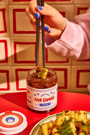 A jar of "MAMA TEAV'S -ORIGINAL -HOT GARLIC CHILI CRISP" with a white label featuring blue and red text and decorative patterns. The jar contains a dark-colored garlic mixture made with organic California garlic. The label highlights the product as "Original" and shows a net weight of 13.7 oz (390g).