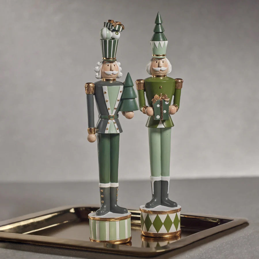 Two decorative ZODAX Nutcracker Soldiers with Gift Boxes stand on a metallic tray. Each figure wears a tall hat and a uniform in shades of green, adorned with intricate details. The product description emphasizes the neutral-toned background, which allows these festive figures to stand out in the foreground.