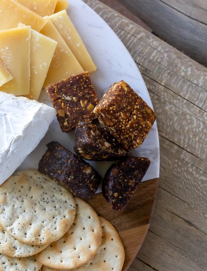 A close-up of a charcuterie board featuring slices of hard yellow cheese, brie cheese, gluten-free FICHI - FIG AND ALMOND BITES from the brand FICHI, and round poppy seed crackers, all beautifully arranged on a white marble and wood serving surface. The board is set on a rustic wooden table.