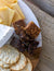 A close-up of a charcuterie board featuring slices of hard yellow cheese, brie cheese, gluten-free FICHI - FIG AND ALMOND BITES from the brand FICHI, and round poppy seed crackers, all beautifully arranged on a white marble and wood serving surface. The board is set on a rustic wooden table.