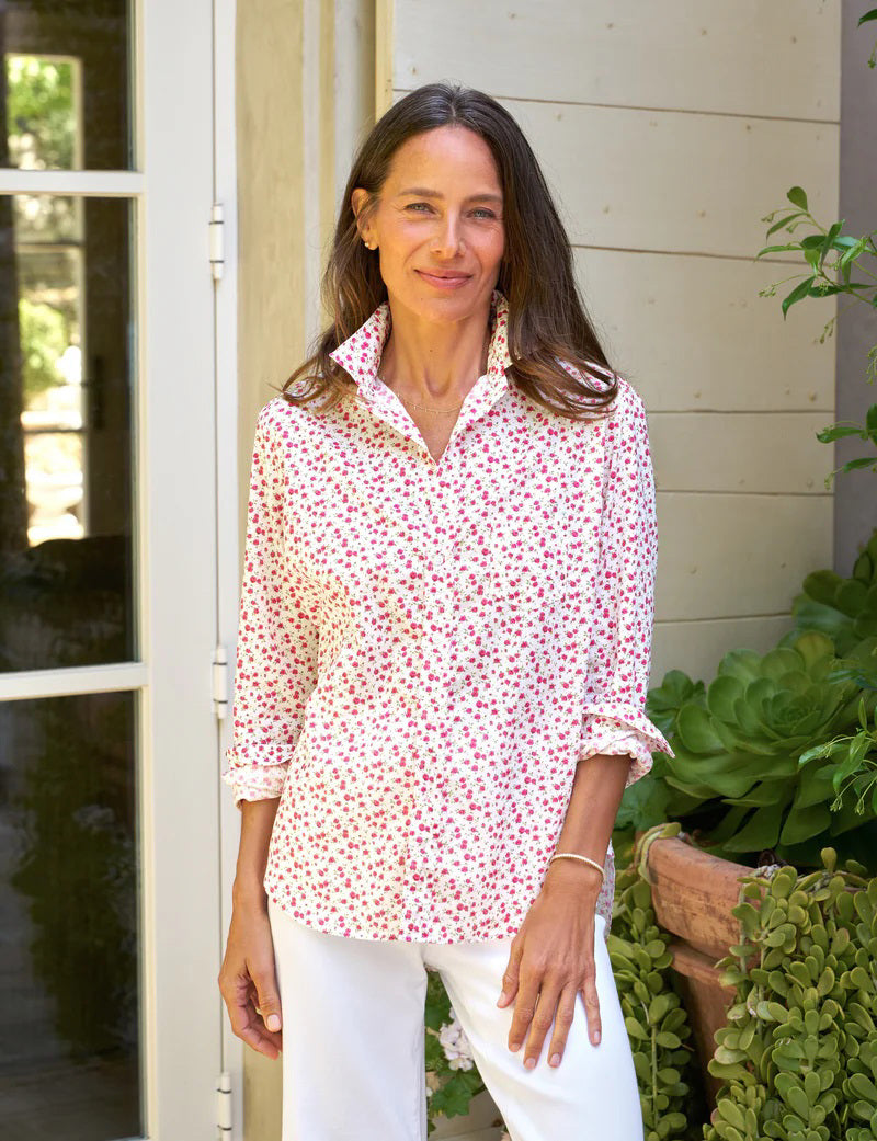 A woman stands outside near a doorway, smiling, her long hair cascading down. She wears FRANK & EILEEN's EILEEN Relaxed Button-Up in Tiny Pink Roses with white pants. Leafy potted plants frame the scene, and the shirt's bust-flattering buttons enhance the natural and peaceful ambiance.