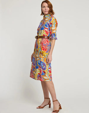 A person with shoulder-length hair is wearing the HINSON WU - TAMRON Long Sleeve Paisley Print Dress with an A-line shape and a brown belt. They are also wearing brown high-heeled sandals and are standing against a plain white background, looking at the camera with a slight smile.