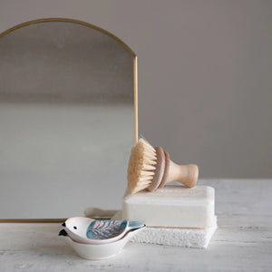 A wooden bristle brush rests on a white soap bar, set before an arched mirror with a brass frame. In the foreground, there is a HAND PAINTED BIRD SHAPED DISH by CREATIVE COOP, featuring a white body and blue wings.