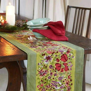 A wooden table features the Jardin Red and Green Table Runner by Couleur Nature, adorned with a lit candle, pinecones, and greenery. Red napkins and silverware are neatly placed next to a light blue bowl on the runner.