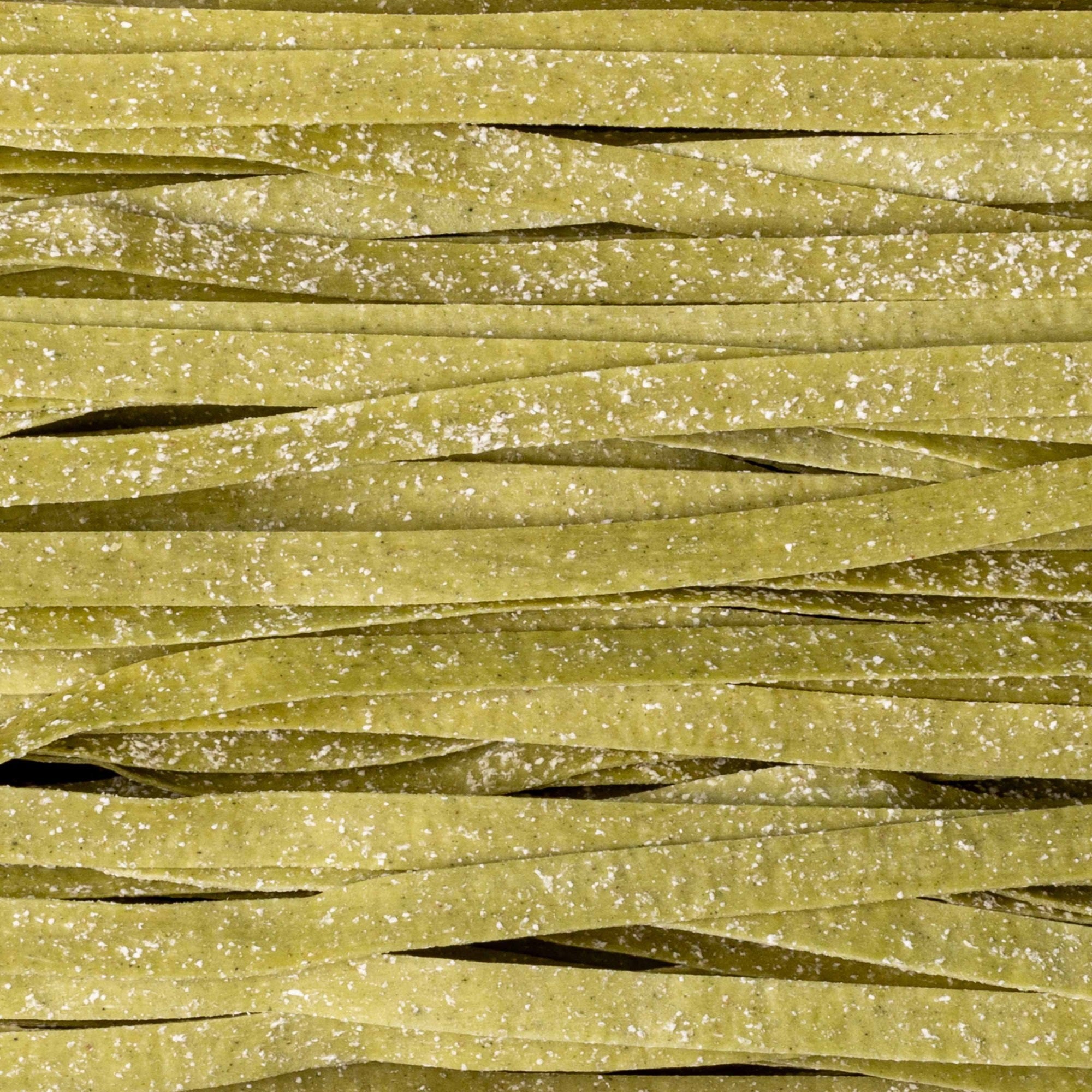 A close-up view of uncooked CIPRIANI - ORGANIC EGG TAGLIARRELLE PASTA WITH SPINACH from ATALANTA CORPORATION. The green-colored egg pasta strands, reminiscent of artisanal pasta, are coated with flour and are arranged in a slightly overlapping manner, showcasing their flat and wide shape.