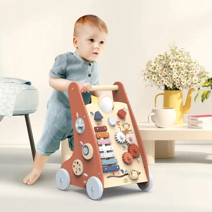 A toddler in a blue outfit energetically pushes the SPEEDY MONKEY - MULTI ACTIVITY WALKER. This toddler learning toy from SPEEDY MONKEY is made from sustainably sourced wood and features interactive elements like a xylophone, gears, and a shape sorter. In the background, a yellow pitcher with flowers, a cup, and books adorn a small table.