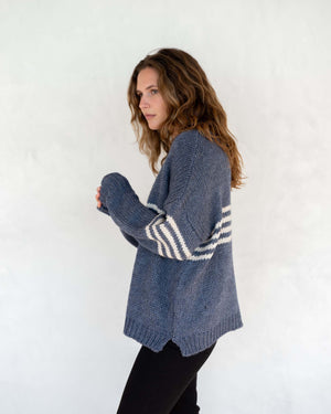 A person with long, wavy brown hair is wearing the MERSEA - SEACOAST STRIPE SWEATER, a loose-fitting, cozy blue knit sweater with white stripes on the sleeves, paired with black pants. They are standing against a plain, light-colored background and facing left, with their hands near their face.