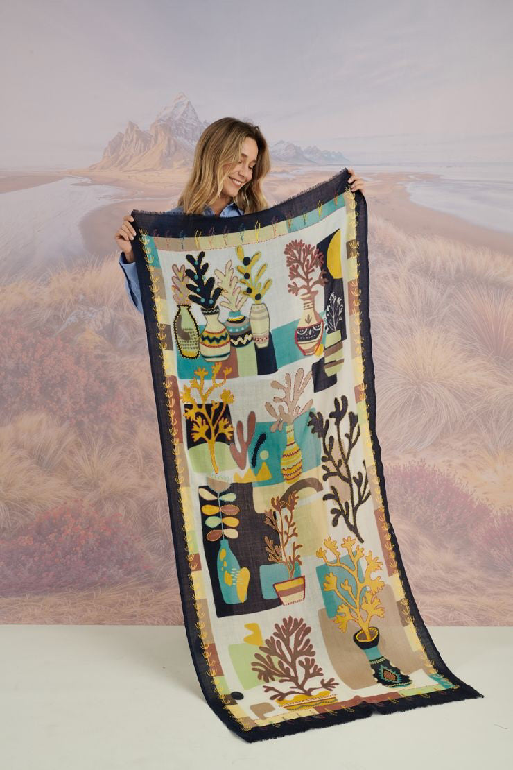 A woman with light brown hair wears a cream blouse and the STORIATIPIC - ANDRES WOOL SCARF by STORIATIPIC, elegantly hand-embroidered with green, yellow, red, and blue patterns. She gazes to the side against a scenic background.