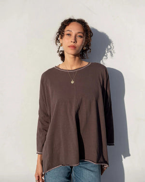 A person stands against a plain white wall, looking at the camera. They have curly hair pulled back and are wearing a loose-fitting, dark gray MERSEA Catalina Slub Long Sleeve Tee by MER SEA with 3/4 length sleeves, blue jeans, and a gold necklace with a circular pendant. Their shadow is visible on the wall behind them.