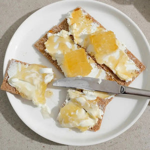 A rectangular tin container labeled "BUSHWICK KITCHEN - 'BEES KNEES' RAW HONEYCOMB" by Bushwick Kitchen. The off-white tin with black and orange text states "Born in Brooklyn, NY" and "Net Wt 7oz (198g)." One side is marked "100% Raw Honeycomb," containing pure, edible honey comb.