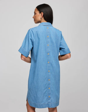 A woman with long dark hair, facing away, wears the HINSON WU - AILEEN Short Sleeve Denim Dress in light blue tencel, featuring a button-down back. She stands against a plain gray background.