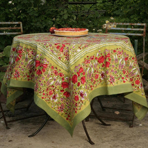 A round outdoor table with a COULEUR NATURE JARDIN RED AND GREEN TABLECLOTH 71X71, featuring floral designs. A fruit tart sits at the center, surrounded by rustic metal chairs akin to Southern France gardens, with lush greenery providing a picturesque backdrop.