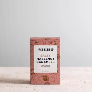 A rectangular pink box with brown floral patterns contains JACOBSEN SALT - SALTY HAZELNUT CARAMELS BOX by JACOBSEN SALT CO. The back of the box features descriptive text about the product's ingredients and origin, highlighting the use of Oregon sea salt and Jefferson hazelnuts for a rich flavor.