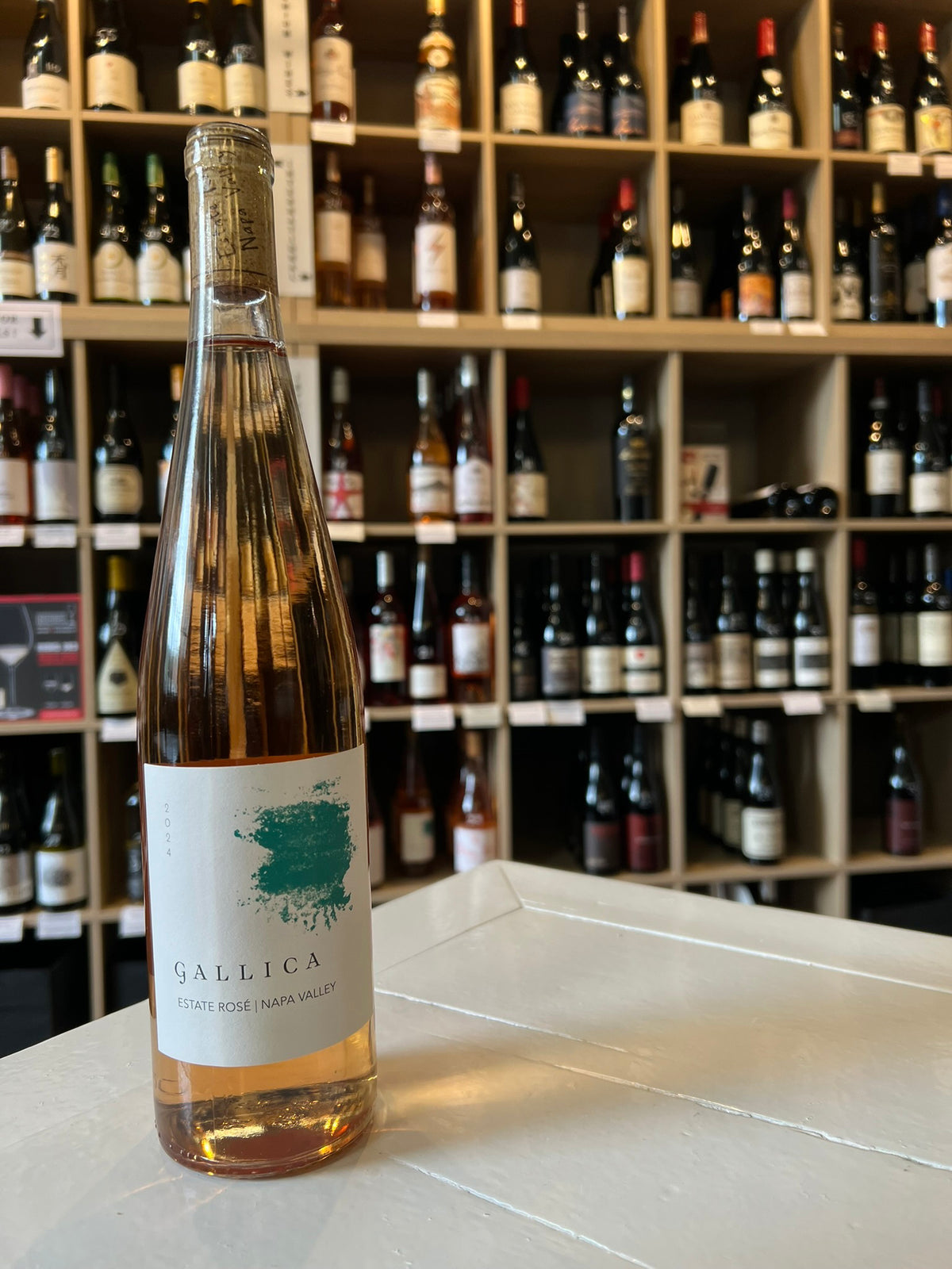 A tall, clear bottle of GALLICA ESTATE ROSÉ 2024 by WINE: JOHNSON BROS./OXFORD ST. gleams golden-pink with a white label and teal splash, topped by cork and gold foil, highlighting the wine’s refined style.