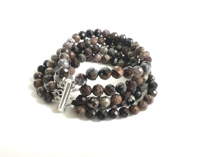 The ANN LIGHTFOOT - FIVE STRAND 6MM FACETED PIETERSITE BRACELET, crafted by Ann Lightfoot, is showcased against a black background. The bracelet features faceted pietersite beads in an array of brown and gray shades, displaying a marbled look, and is secured with a stainless steel clasp.