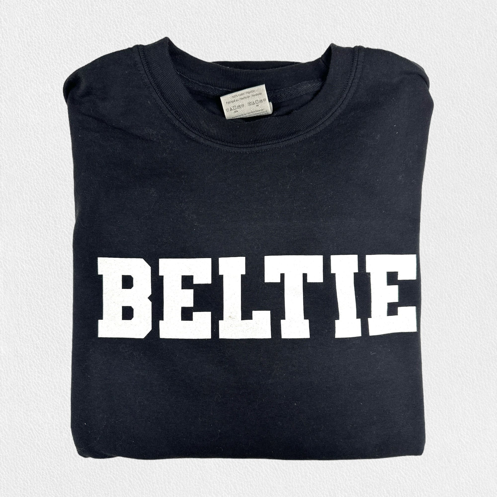 A black long sleeve tee shirt from the Fearrington Lifestyle Collection by Comfort Colors, crafted from premium fabric with the word "BELTIE" printed in large, bold, white capital letters on the front, neatly folded and laid on a white textured surface.