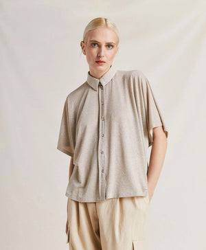 A woman with platinum blonde hair stands with a neutral expression, wearing the MOMONI - BROOKLYN LUREX JERSEY BLOUSE and colorful beaded necklaces. She has her hands in the pockets of her striped trousers with oversize lines. The background is plain and softly lit.